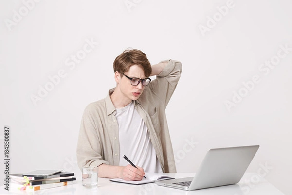 Obraz Portrait of thoughtful young man student wears beige shirt and spectacles looks concentrated and study at the table using laptop computer and notebooks isolated over white background