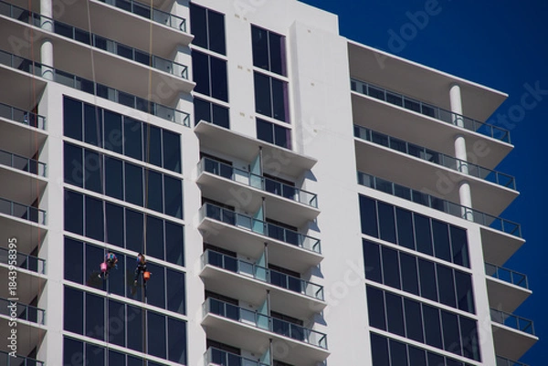 Obraz Modern High-Rise Building With Glass Facade And Rappelling Window cleaners Workers On A Clear Day in St. Petersburg, FL. Expansive glass panels and stacked balconies. Two workers in safety gear descen