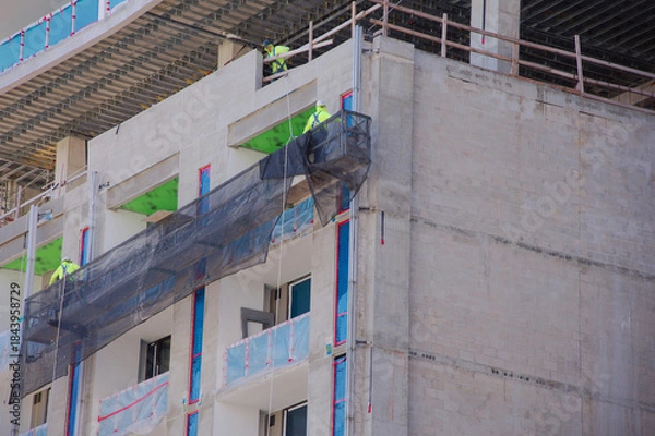 Obraz Construction Workers in safety gear Install Exterior Facade Using Suspended Safety Net Scaffolding on Building Under Construction Site in St. Petersburg, FL. Partially built building, showcasing teamw