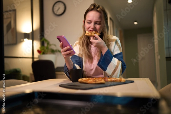 Obraz Female eating pizza while using smartphone at kitchen table