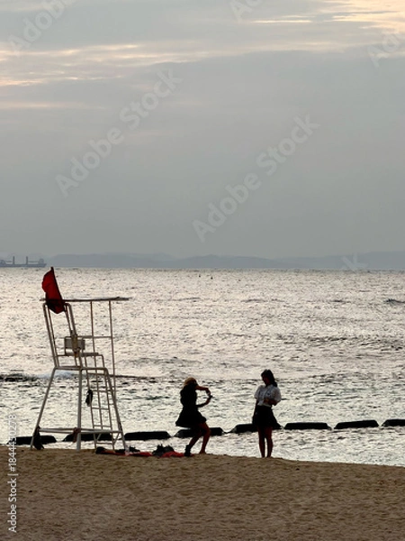 Obraz Two young women taking photos on a sandy beach near a lifeguard tower at sunset