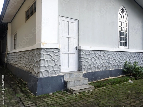 Fototapeta Exterior corner of a traditional building featuring a white door, an arched window, and a gray, textured stone-patterned foundation with mossy brick pavers in the foreground.