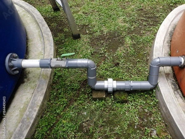 Fototapeta Overhead view of a gray PVC pipe with a green ball valve connecting the base of a blue water tank to an orange water tank, set on a grassy area with concrete curbs.