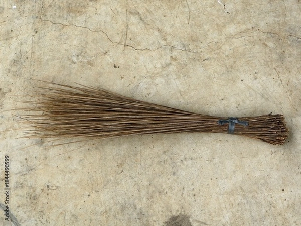Fototapeta Traditional Indonesian or Southeast Asian broom Sapu Lidi made from bundled coconut palm leaf ribs, resting on a textured, cracked concrete floor. Rustic and eco-friendly cleaning tool.