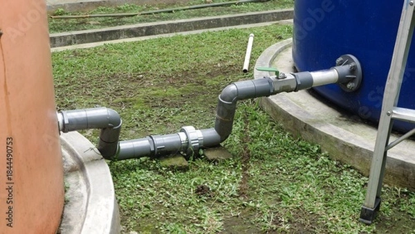 Fototapeta A gray PVC pipe connection, including a green ball valve, linking two large outdoor plastic water storage tanks orange and blue on a grassy area with concrete curbs.
