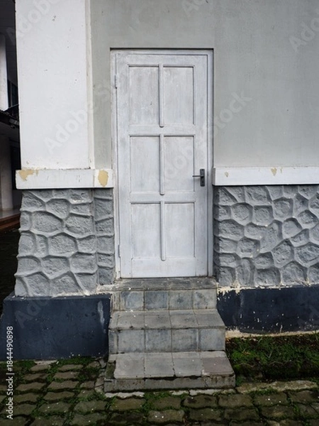 Fototapeta A quiet, weathered white wooden six-panel door framed by gray walls and a decorative stone-patterned foundation. Low stone steps lead up from mossy brick paving.