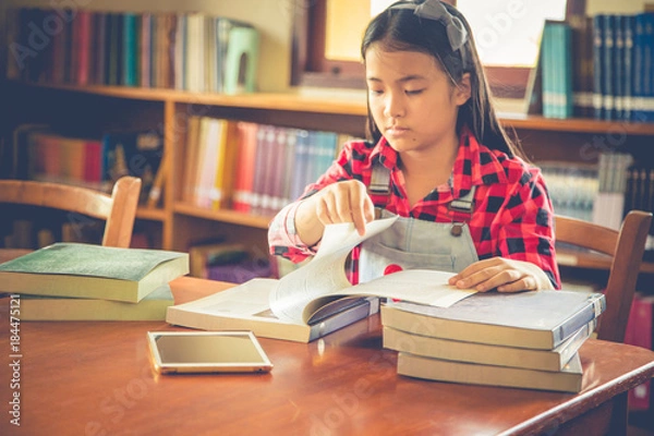 Fototapeta Girl is using tablet while sitting in library and reading a book