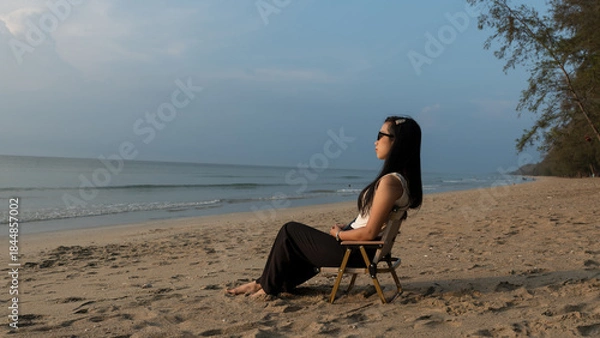 Obraz Woman sitting on a beach chair facing the sea under soft golden sunset light.