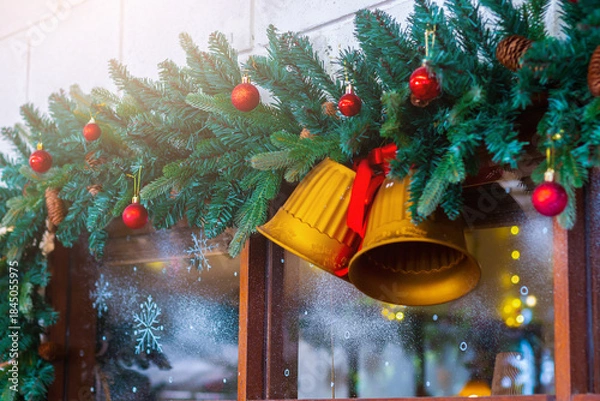 Fototapeta Festive Christmas decoration with evergreen garland, golden bells, and red ornaments hanging above a window, creating a warm holiday atmosphere.