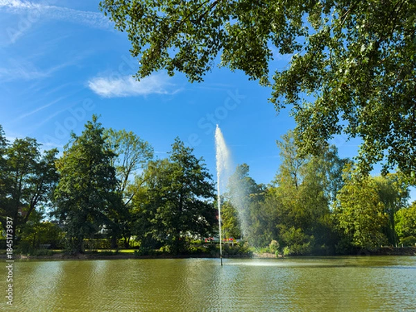 Obraz Fountain sprays water into the clear blue sky above lush greenery and a serene pond on a sunny afternoon in a tranquil park setting