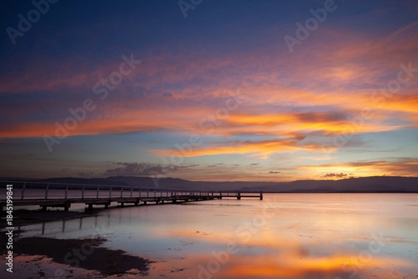 Fototapeta Boonerah Point jetty under a beautifully vibrant sky looking west to the mountains on Lake Illawarra, New South Wales, Australia.