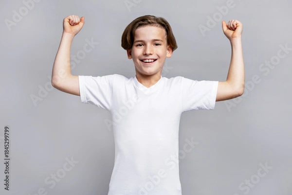 Obraz Young boy with happy face celebrating victory. Child expressing success and achievement. Kid showing strength and positive emotion in bright studio on grey isolated background