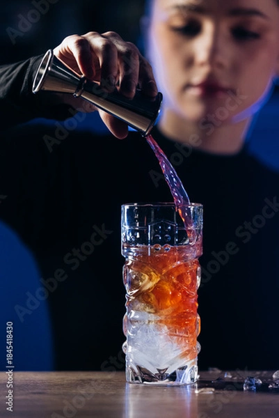 Obraz Female bartender skillfully pouring a vibrant cocktail into a glass filled with ice, showcasing mixology techniques in a modern bar environment with ambient lighting