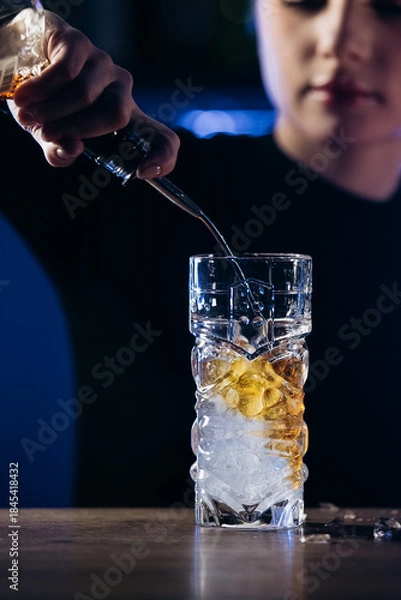 Obraz Bartender pouring a drink into a glass filled with ice, showcasing cocktail preparation skills in a vibrant bar environment with atmospheric lighting and modern decor