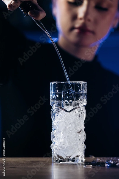 Obraz Female bartender pouring liquid into a glass filled with ice, showcasing cocktail preparation skills in a dimly lit bar environment with vibrant colors