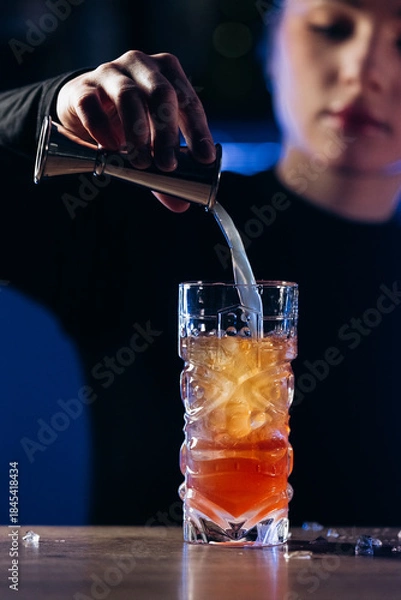 Obraz Bartender pouring a colorful cocktail into a glass filled with ice, showcasing mixology skills and vibrant drink presentation in a stylish bar environment
