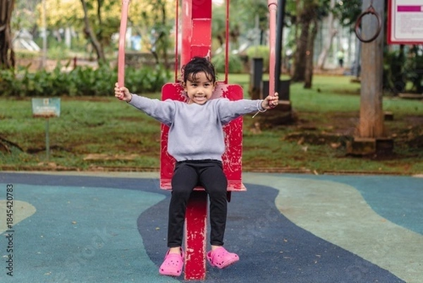 Obraz A little Indonesian girl exercising outdoors in the park, trying out a shoulder pull exercise machine