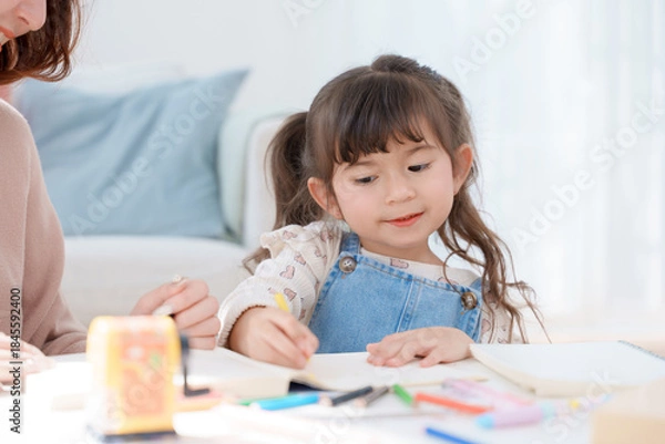 Fototapeta Cheerful Asian mother and little daughter enjoying creative activity together at a bright, cozy white table in the living room. Focus on happy family bonding, learning, and childhood development.