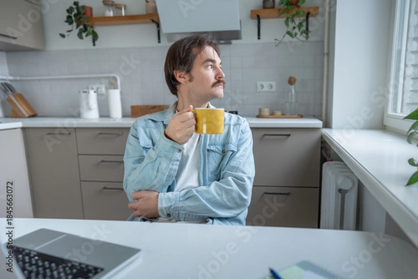 Fototapeta Calm reflective man holding mug of coffee in home kitchen, looking away deep in thought. Pensive young man with cup in hand taking break at table, gazing out window. Inner dialogue, decision making.