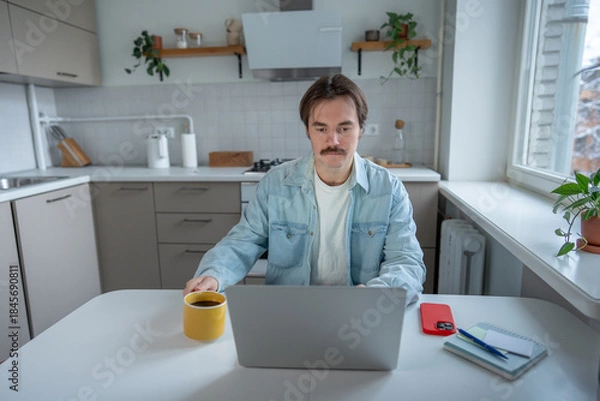 Fototapeta Focused serious man remote working with laptop at kitchen table holding cup coffee, checking plan for day. Morning productivity, balancing work and home comfort, self-discipline, daily work routine.
