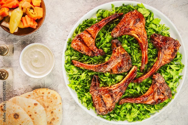 Obraz Middle Eastern traditional dish with grilled juicy lamb chops/ribs placed on bed of fresh chopped parsley. Served with tahini sauce, Arabic bread and pickles.