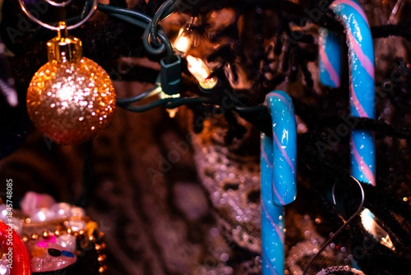 Fototapeta Close up of a light blue candy cane hanging on a tree as holiday decor. The tree is lit with lights and has a shiny gold ornament hanging close to the candy decor. 
