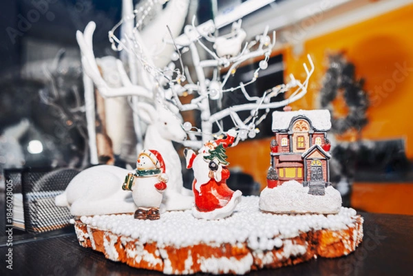 Obraz Festive Christmas scene features white reindeer, snowman, Santa, and fairy house nestled in snowy decor. Warm lights and a blurred wreath enhance the cozy, celebratory holiday atmosphere