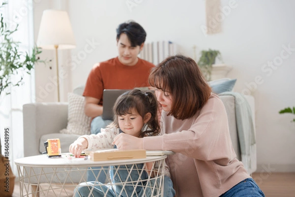 Fototapeta A cheerful Asian mother helps her young daughter do homework and color a book at home, while the father works on a laptop in the background. A cozy, bright, and modern family lifestyle.