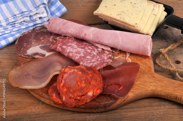 Obraz Assortiment de charcuteries sur une planche à découper en bois avec du fromage à raclette tranché dans un poêlon