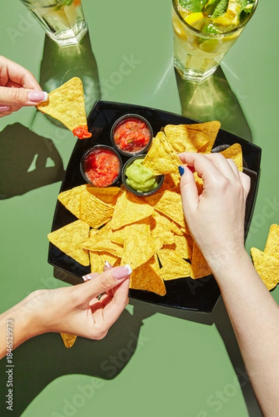 Obraz Female hands holding nachos corn chips and dipping them in salcha and guacamole sauces, glasses with lemonade are standing nearby. Mexican cuisine concept