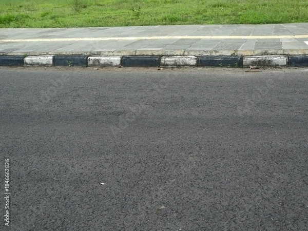 Fototapeta Low-angle view of an asphalt road meeting a sidewalk with black and white painted curb blocks, bordered by a green grass verge above.