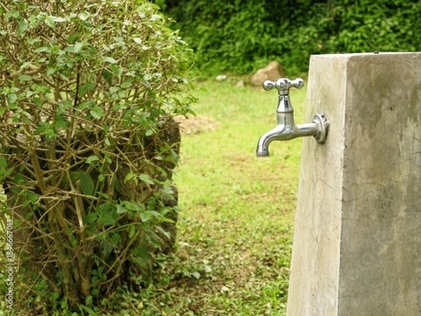 Fototapeta A bright chrome faucet installed on a rough concrete pillar in a lush green outdoor setting, with a dense green bush on the left.