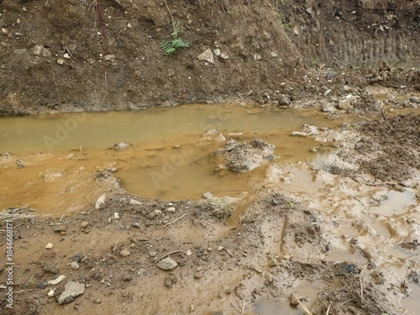 Fototapeta A very close-up detail of a muddy ditch filled with brown, opaque water and large clumps of churned earth, rocks, and visible tire tracks.