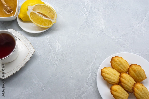 Fototapeta Madeleine cookies on a white plate with lemon, honey, and a cup of tea against a light background. Space for text.