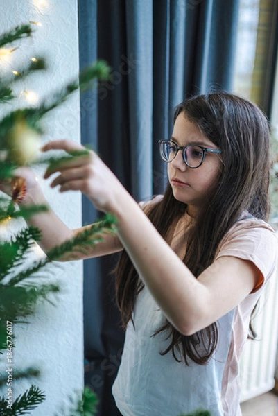 Fototapeta Teenage girl in glasses and T-shirt decorating Christmas tree at home, cozy holiday family moment