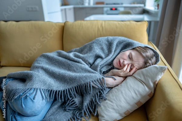 Fototapeta Sick woman wrapped in blanket lying on sofa at home, experiencing chills, body aches, fatigue. Female feeling unwell during cold winter season touching face with closed eyes suffering headache