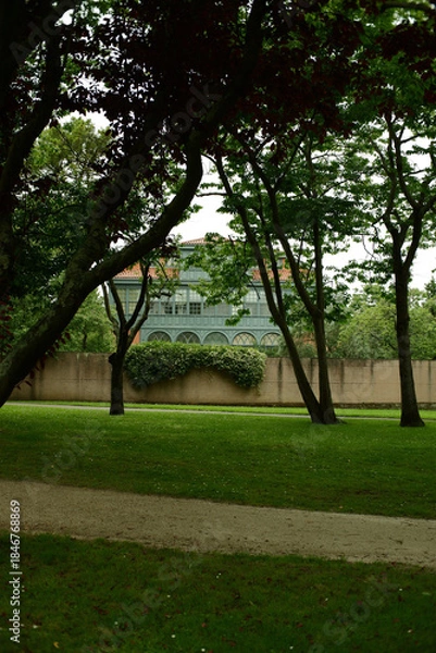 Fototapeta Park Ferrera in Aviles on a summer overcast day, featuring green lawns, trees, and historic buildings. Empty urban park with a calm and serene atmosphere