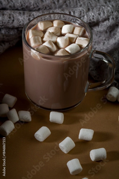 Fototapeta Warm drink with marshmallows in a glass mug on a table with scattered marshmallows around it in the evening light