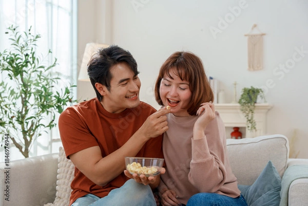 Fototapeta Happy young Asian couple eating popcorn together, man feeding woman in bright living room. They are relaxing and bonding while sitting on the sofa at home.