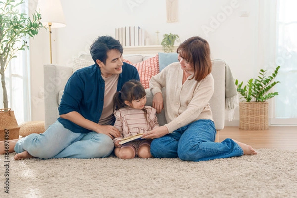 Fototapeta Asian parents and cute daughter sit on a plush, sunny rug, reading a storybook together. Happy family bonding through learning in the cozy living room.