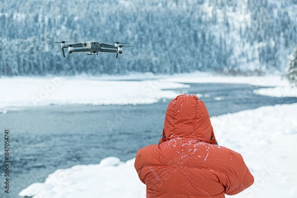 Fototapeta Flying drone with operator in orange jacket against winter background with river and forest