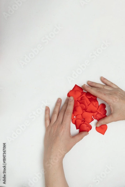 Obraz Two hands are sorting through a pile of small red hearts on a plain white surface. This scene shows preparation for Valentine's Day themed digital content.