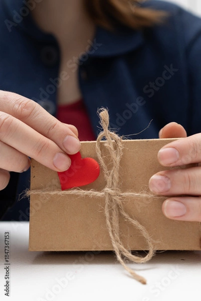 Obraz Hands decorate a simple brown gift box. A small red heart is added to the box. The box is also tied with natural brown string. Presented for Valentine's Day on a white surface.