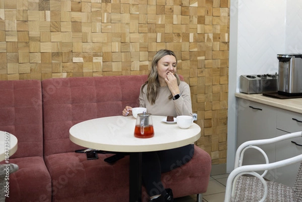 Obraz Young woman in a beige shirt sits at a round table in a cafe, eating dessert and drinking tea. She smiles and looks pleased while enjoying her afternoon treat.