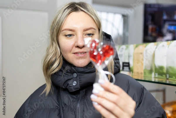 Obraz A blonde woman in a black puffer jacket holds and looks at a red, heart-shaped lollipop wrapped in plastic. She is inside a store during the day.