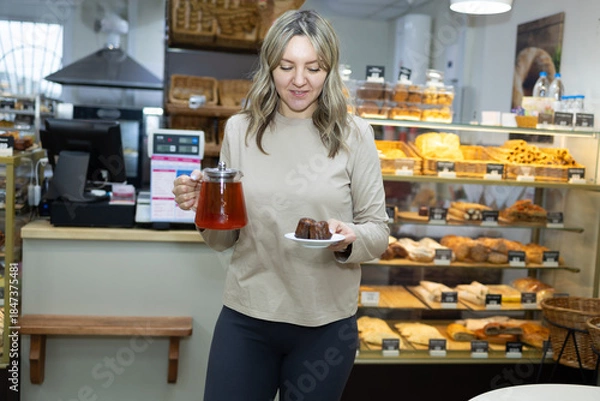 Obraz A blonde woman inside a bakery holds a plate with two caneles and a teapot with red liquid. A display case filled with pastries sits behind her at the bakery.