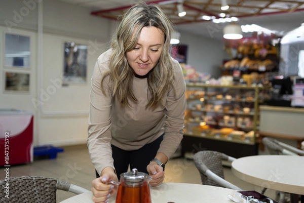 Obraz A woman in a tan long sleeve shirt prepares to serve tea at a table inside a cafe. A glass display case filled with desserts is behind her. There are empty tables and chairs around her.