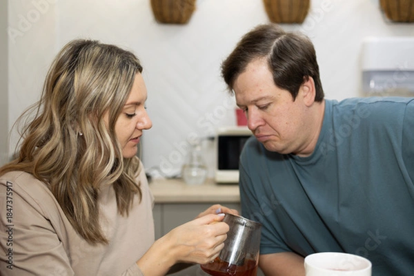 Obraz In a kitchen setting, a woman pours tea from a glass pitcher into a white mug for her husband. He looks down at the tea, waiting. They are standing near a counter.