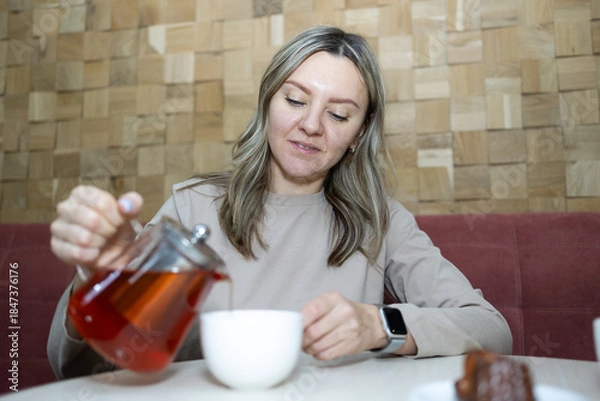 Obraz A young woman with blonde hair pours red tea from a clear teapot into a white cup. She sits at a table in a cafe with a wood block wall and a dark red banquette.