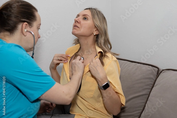 Obraz A healthcare professional examines a patient sitting on a couch in a living room. The doctor uses a stethoscope to listen to the patient's heart. The patient appears attentive during the check-up.